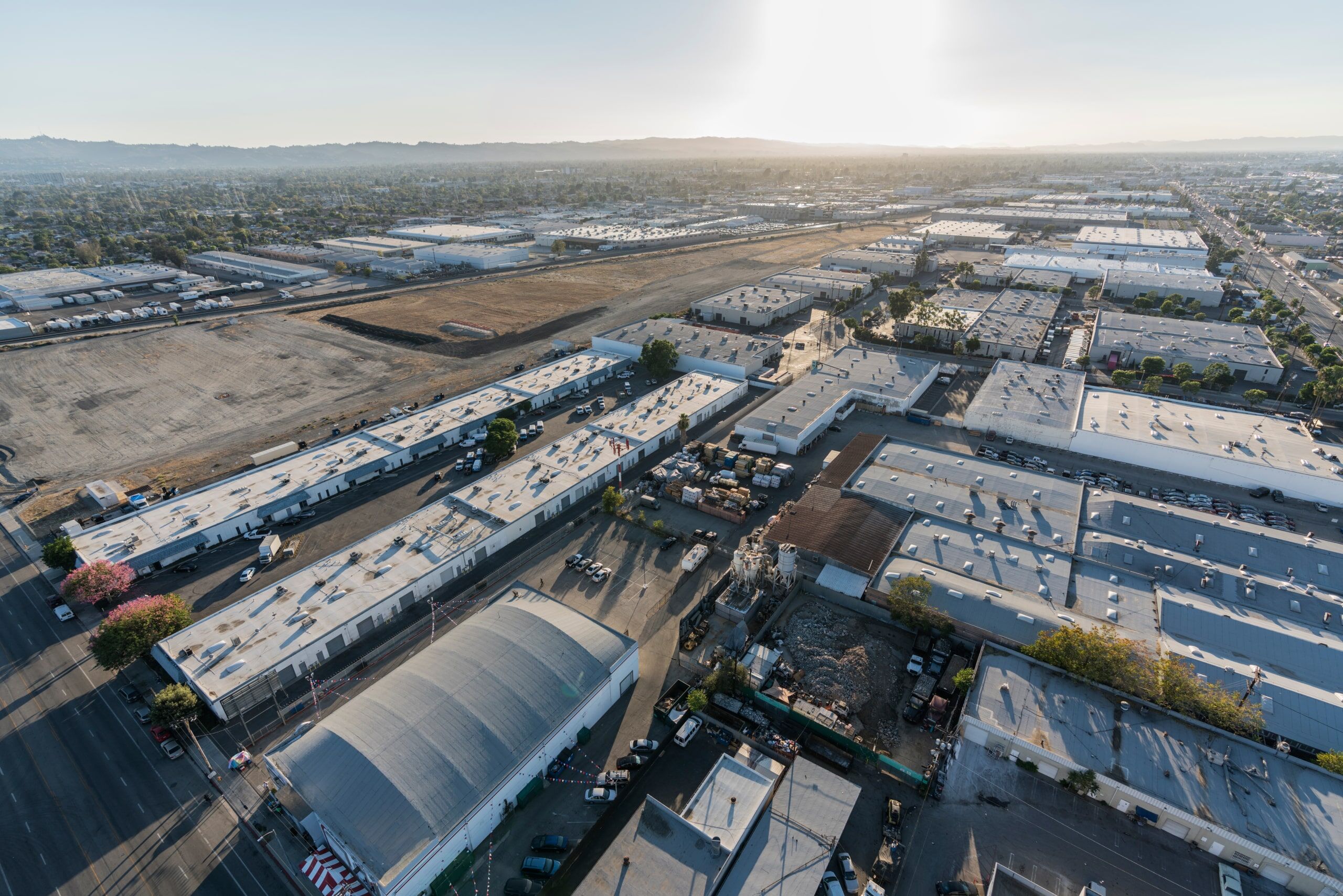 Aerial view of an industrial area with large warehouses, paved roads, parked vehicles, and open lots under a hazy sky with the sun low on the horizon.