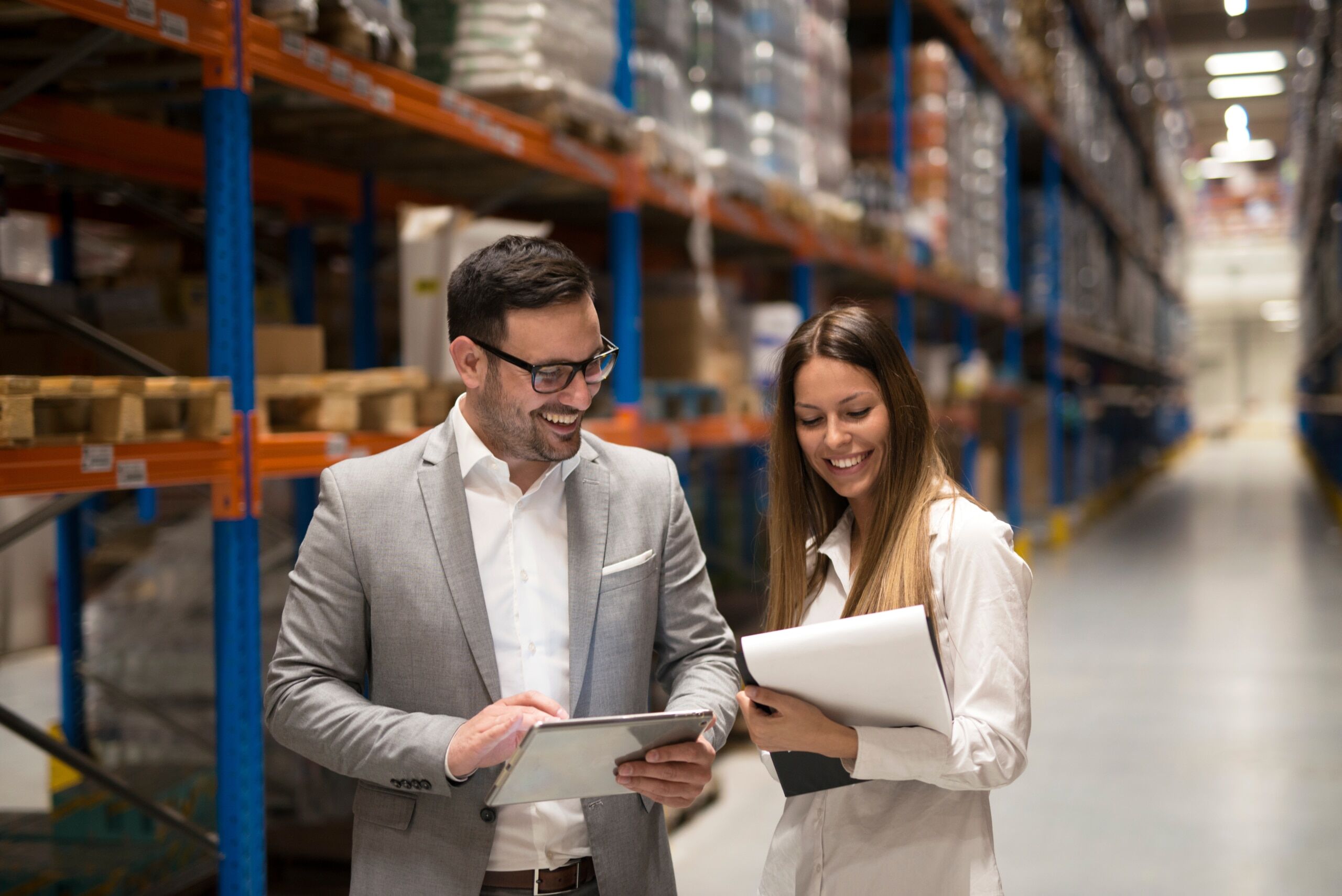 Two people stand in a warehouse aisle, smiling and looking at documents and a tablet. Shelves stocked with boxes and items are visible in the background.