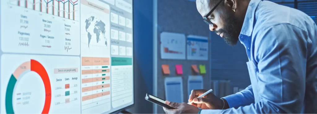 A man analyzes data on a large screen displaying charts, graphs, and maps while making notes on a tablet in an office setting.