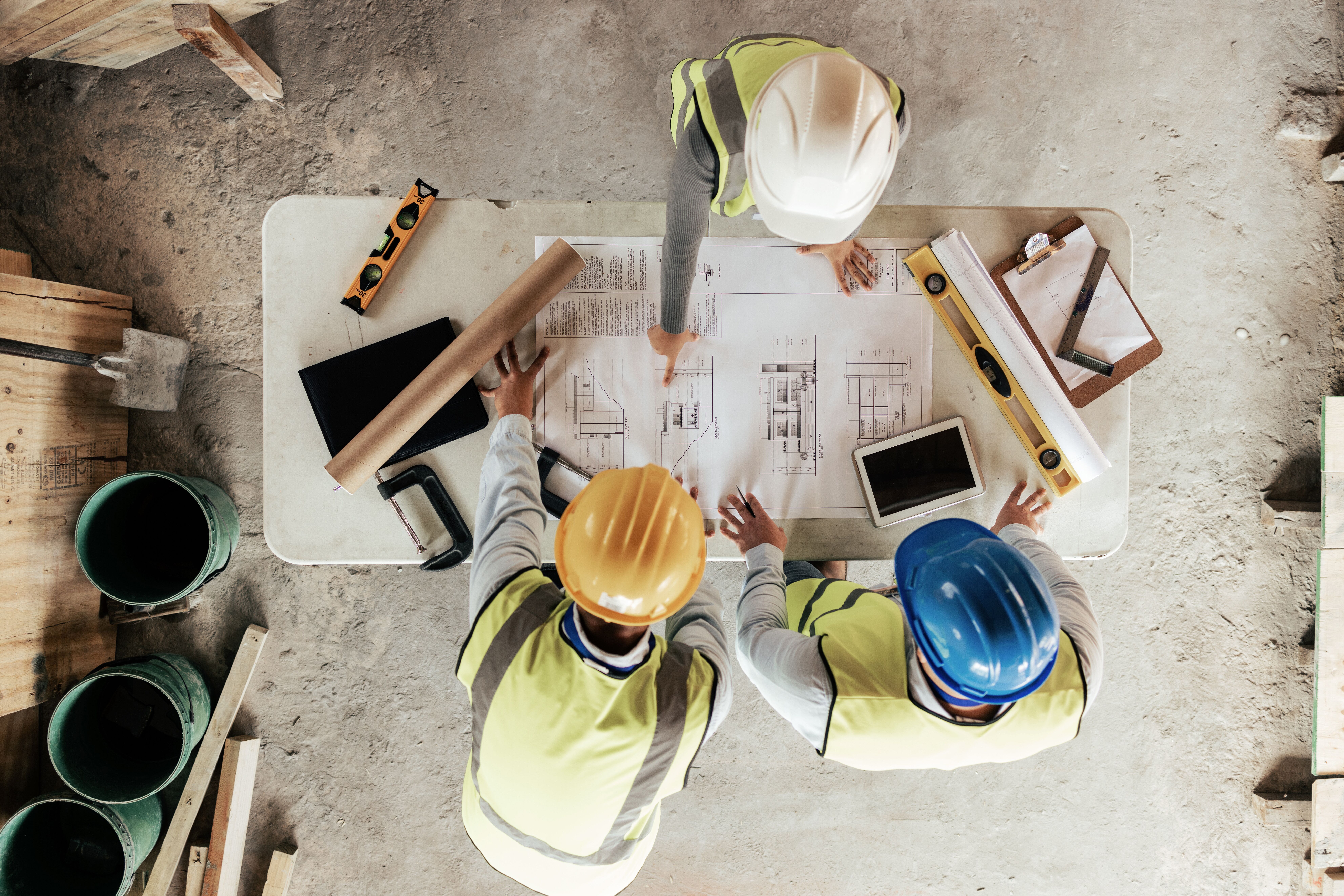 Three construction workers in safety gear review architectural blueprints on a table, surrounded by tools, a laptop, a tablet, and building materials.