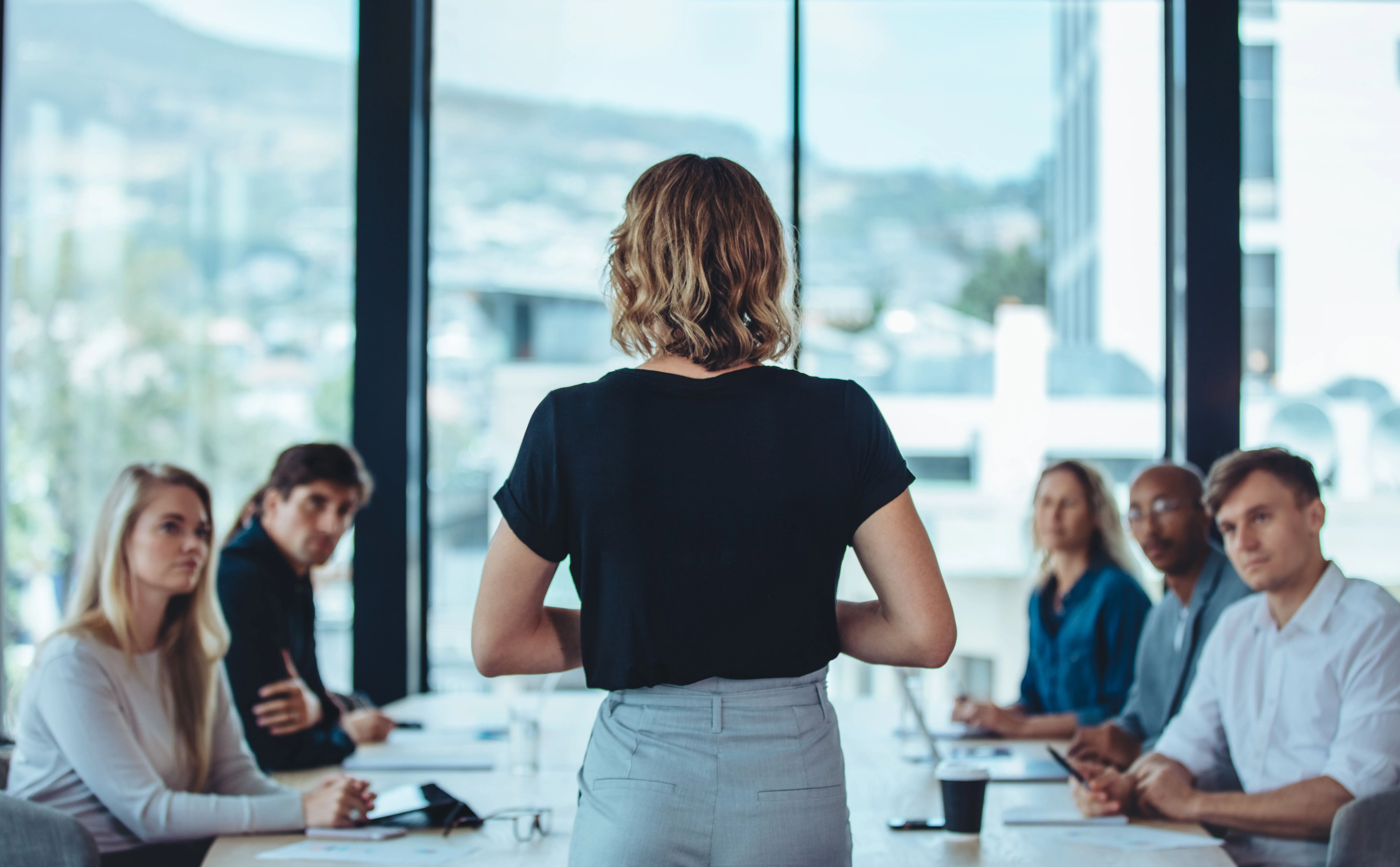 A woman stands and speaks to a group of five people seated at a conference table in a modern office setting.