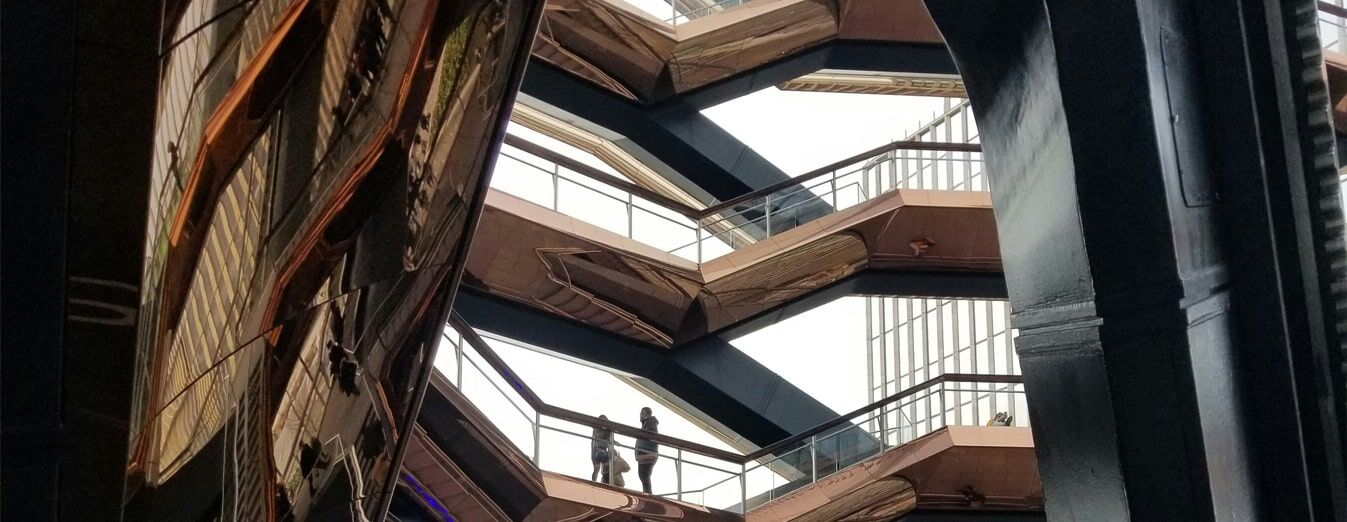A person stands on an upper walkway of a modern, zigzagging outdoor staircase structure with geometric metal railings.