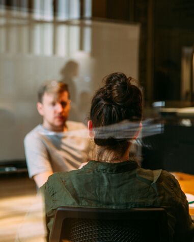 Two people sit across from each other at a table in an office setting, with one person facing the camera and a blurred whiteboard in the background.