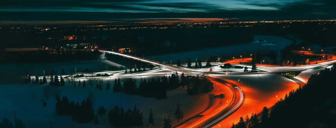 Aerial view of a snowy city intersection at dusk, with car headlights and taillights creating light trails on the roads.