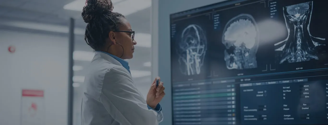 A doctor in a white lab coat examines brain and neck MRI scans displayed on a large monitor in a medical setting.