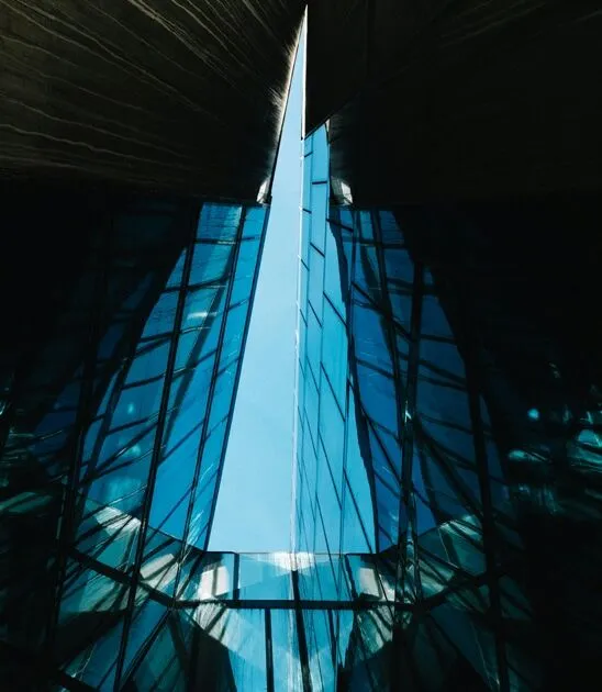 View looking up at the sharp, angular architecture of a modern glass building against the sky, framed by dark structural elements.
