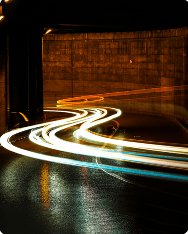 Long-exposure photo of light trails from moving vehicles on a wet, curving road at night under an overpass.
