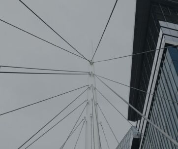 View looking up at the cables of a modern suspension bridge between two glass office buildings against a gray sky.