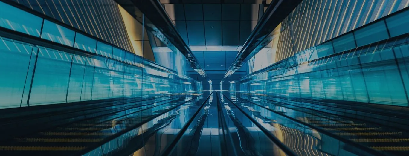 A modern, empty escalator tunnel with blue lighting and reflective glass walls, creating a symmetrical, futuristic effect.