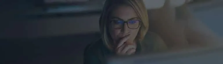 A woman wearing glasses looks at a computer screen with a thoughtful expression, resting her hand on her chin.