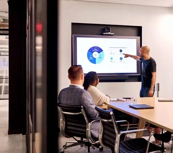 A man stands and points at a presentation slide with charts on a screen while two seated colleagues watch in a modern conference room.
