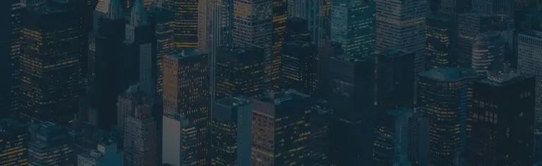 Aerial view of a cityscape with numerous tall buildings and skyscrapers, many of which have their lights on in the windows during dusk or nighttime.