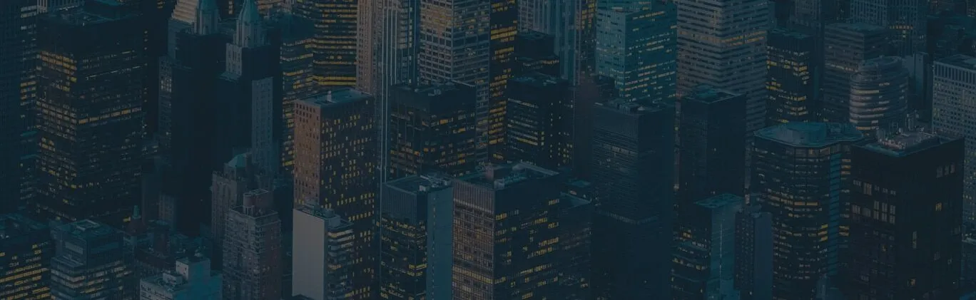 Aerial view of a cityscape with numerous tall buildings and skyscrapers, many of which have their lights on in the windows during dusk or nighttime.