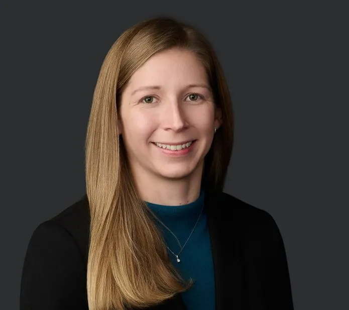 A woman with straight, long blonde hair wearing a black blazer and teal top smiles at the camera against a plain dark background.