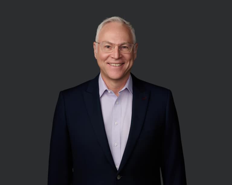 A middle-aged man with short gray hair and glasses smiles at the camera, wearing a dark blazer and light purple shirt, against a plain dark background.
