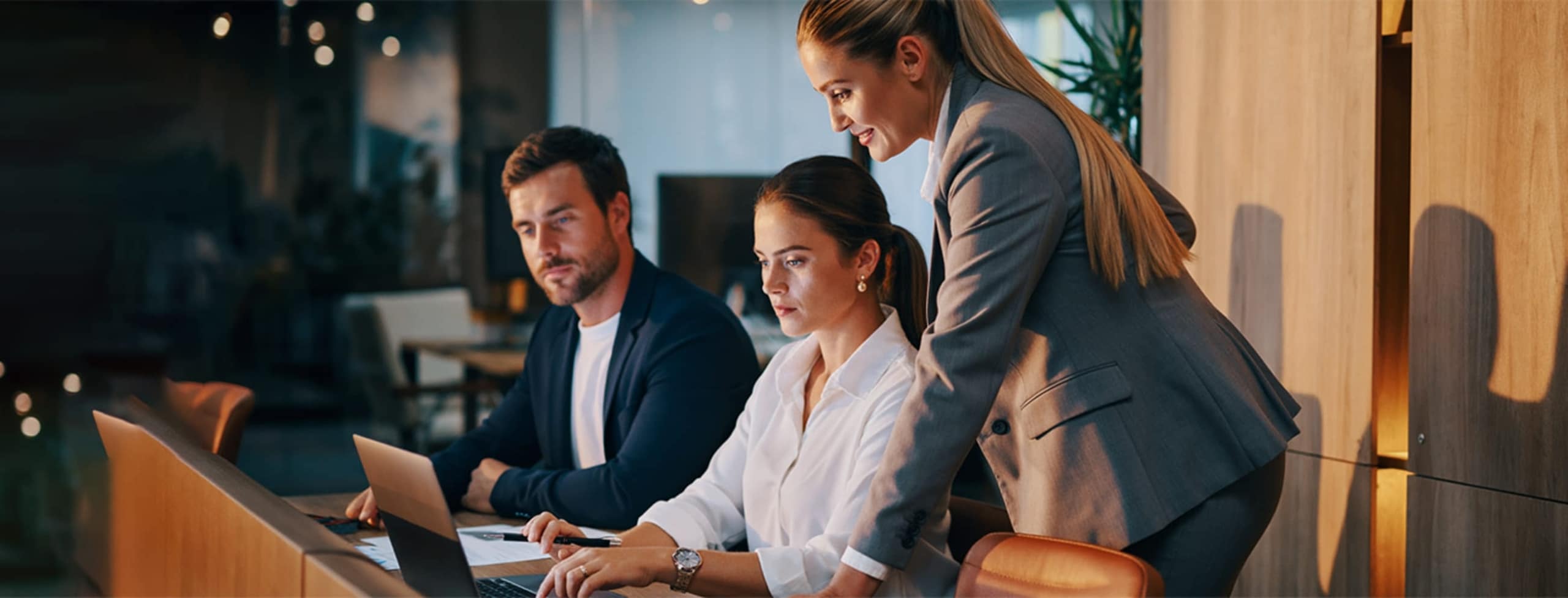 Three professionals in business attire work together at a desk with laptops, reviewing information and discussing in a modern office setting.