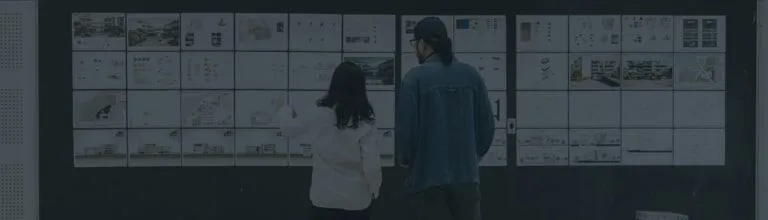 Two people stand in front of a wall covered with various printed diagrams, charts, and architectural plans, examining the materials closely.