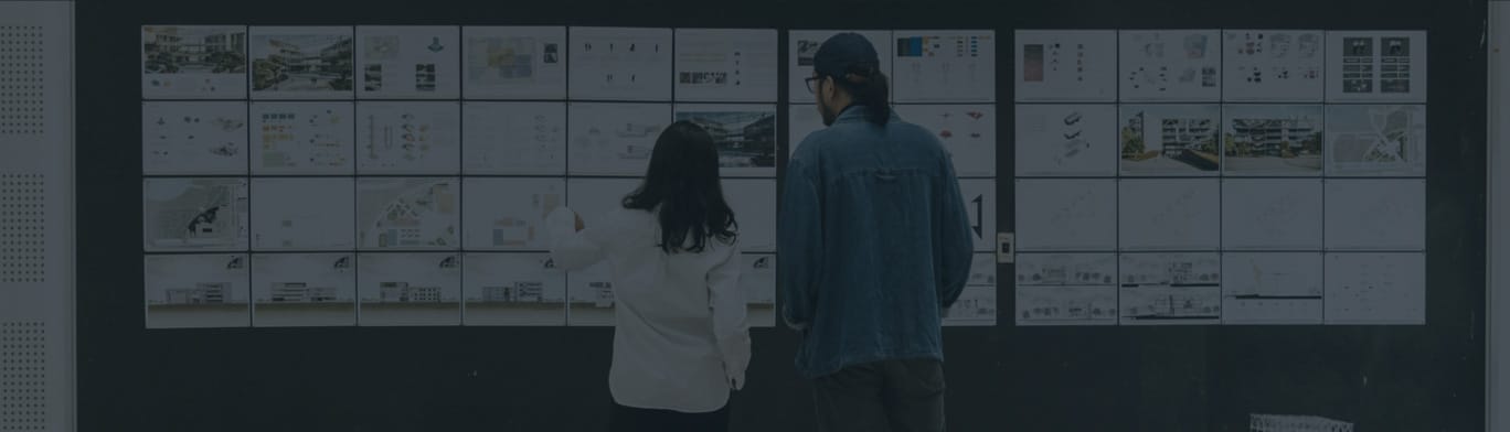 Two people stand in front of a wall covered with various printed diagrams, charts, and architectural plans, examining the materials closely.