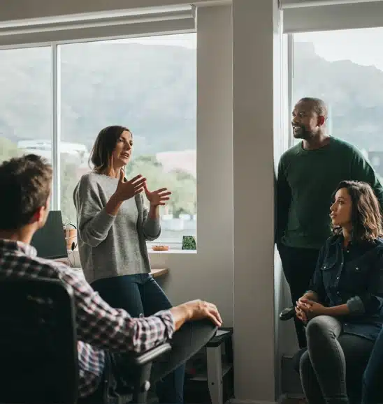 Four people sit and one stands in a bright office, having a discussion near large windows with a view of hills outside.
