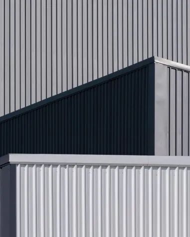 A close-up view of the exterior of a modern industrial building with overlapping geometric shapes and vertical metal siding in shades of grey.