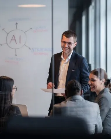 A man in a suit presents to three colleagues in a conference room with a whiteboard showing "AI" and related terms.