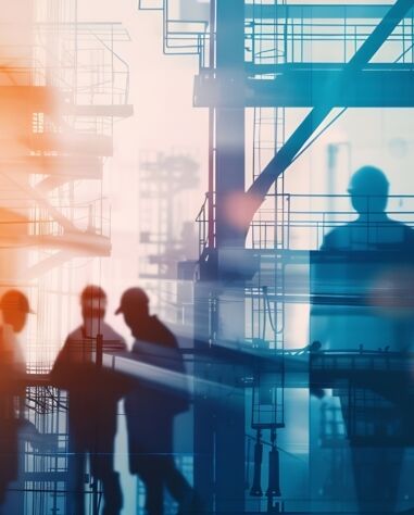 Silhouettes of workers wearing hard hats stand and walk at an industrial construction site with metal scaffolding and framework in the background.