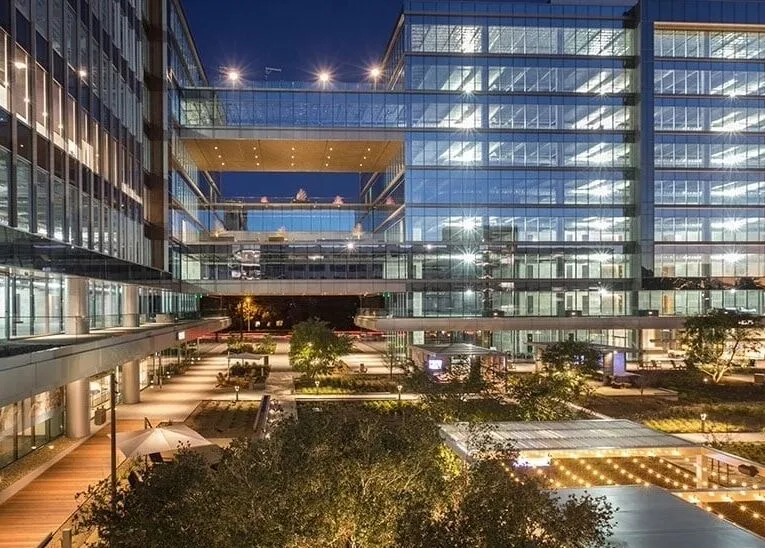 Modern glass office buildings with illuminated windows and outdoor courtyard, photographed at dusk.