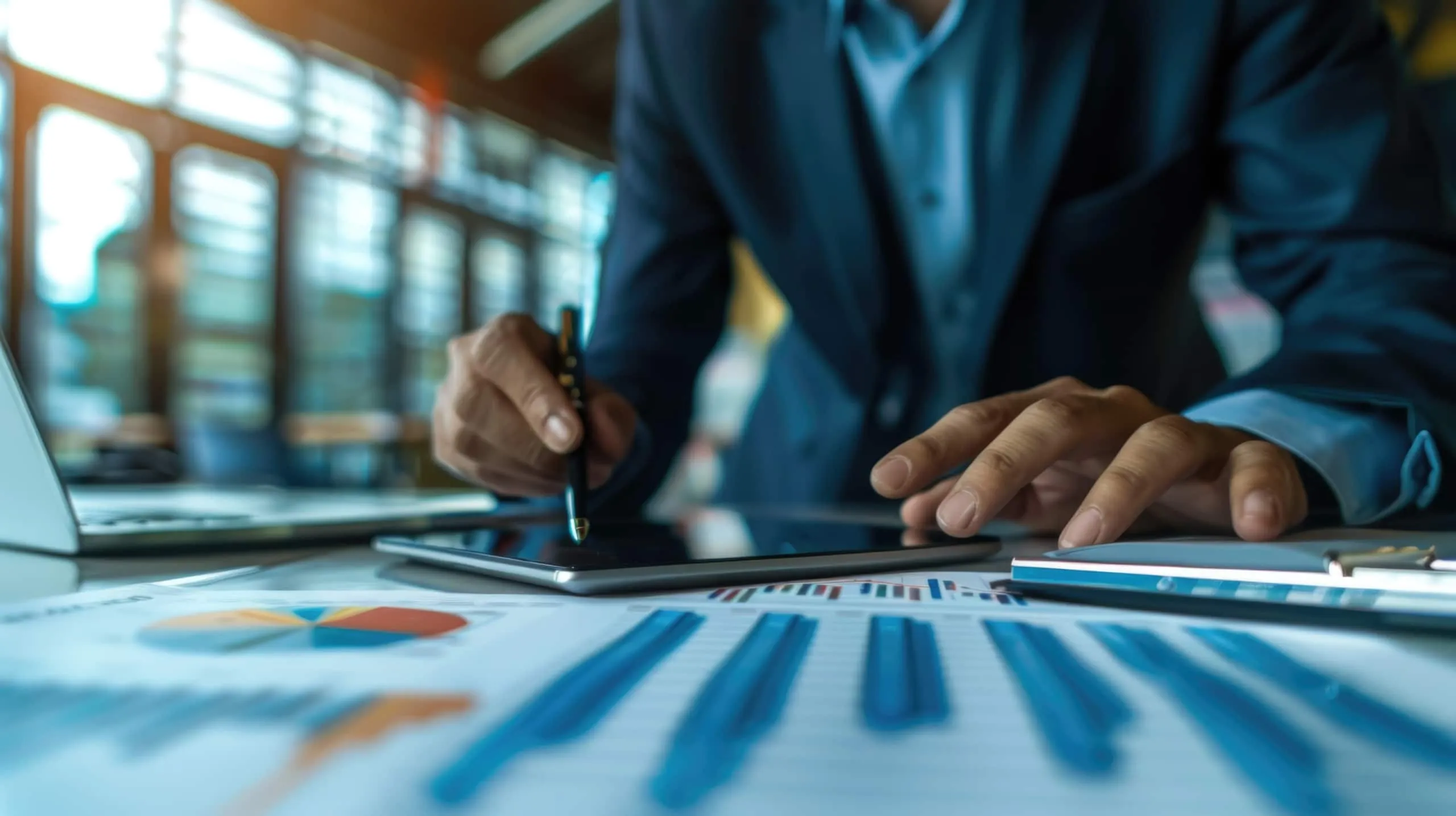 A person in a suit uses a tablet with one hand while holding a stylus, reviewing a due diligence checklist as charts and graphs are visible on the desk in the foreground.