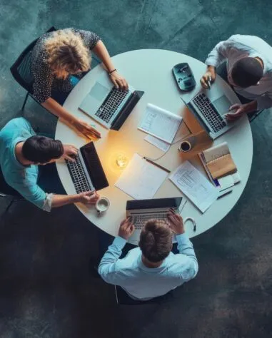 Four people conducting due diligence as they work on laptops around a round table, surrounded by documents, notebooks, and coffee cups, viewed from above.