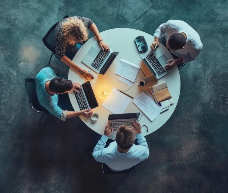 Four people conducting due diligence as they work on laptops around a round table, surrounded by documents, notebooks, and coffee cups, viewed from above.