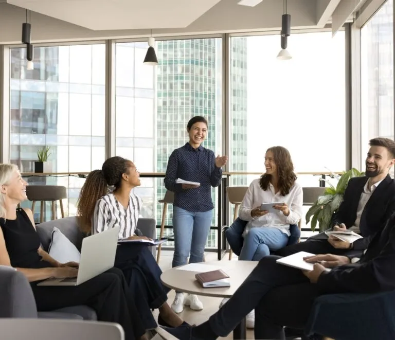 Six people sit in a modern office lounge, listening to a standing woman who appears to be presenting or leading a discussion. Laptops and notebooks are visible. Large windows show city buildings.