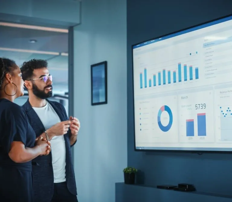Two people stand in front of a large screen displaying charts and graphs, discussing data visualizations in a modern office setting.