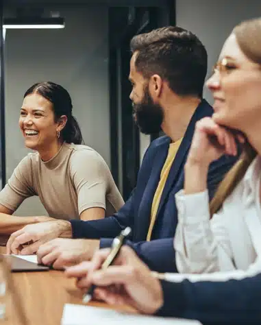 Three people sit at a conference table, engaged in conversation; one woman is smiling and laughing while the others listen attentively.