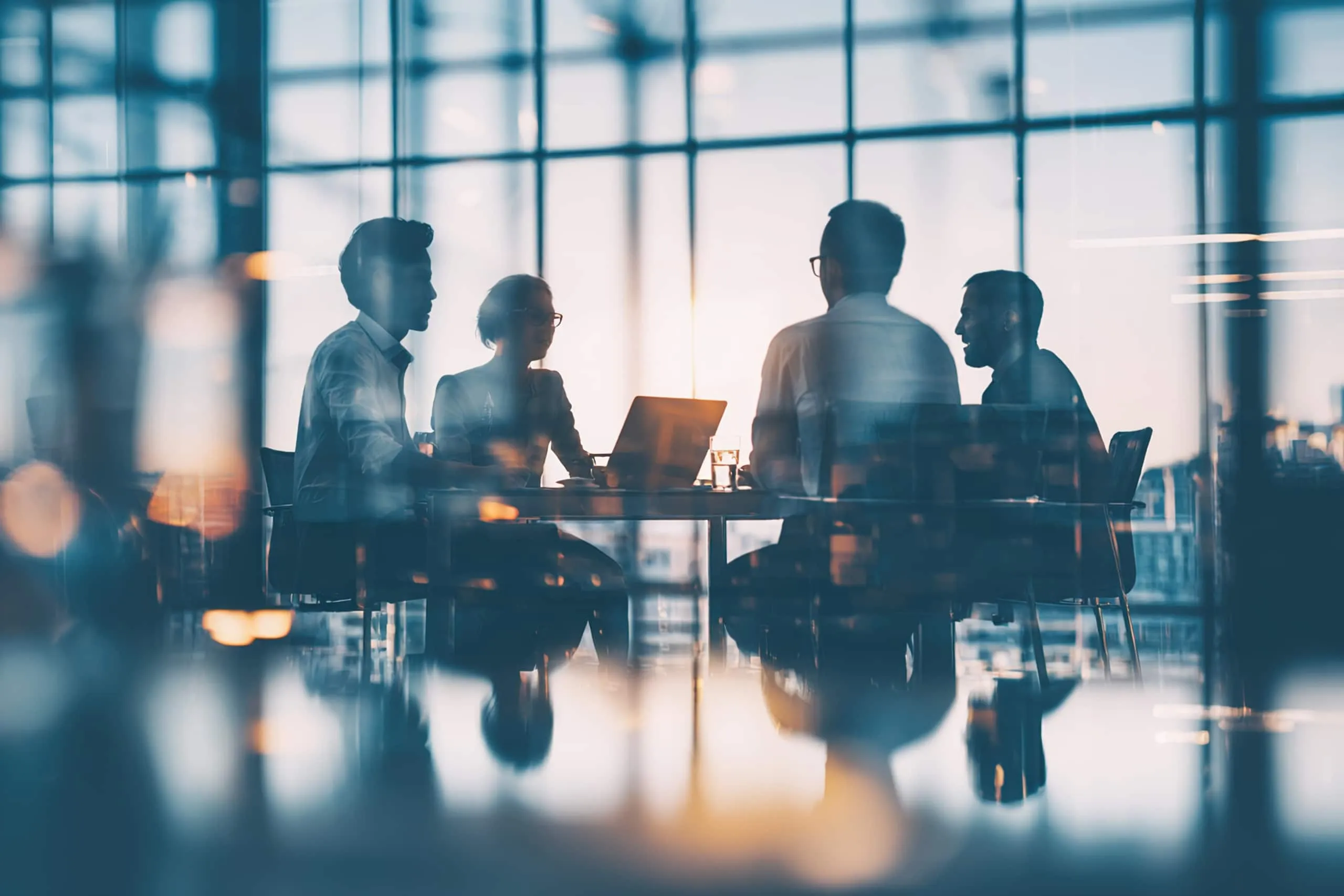 Four people sit at a table in an office meeting room with large windows, silhouetted by natural light and reflections.