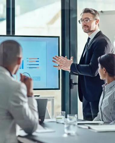 A man in a suit gives a presentation to two colleagues in an office, displaying a bar chart on a large monitor.
