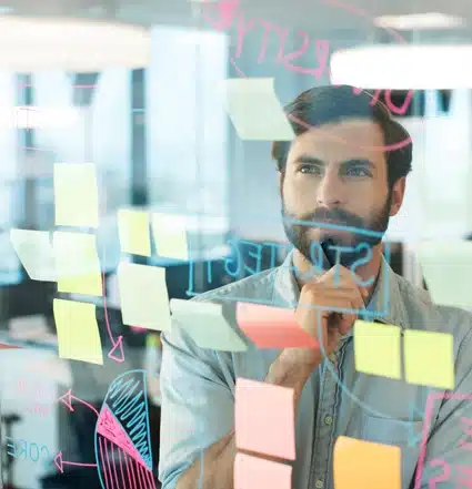 A man stands behind a glass wall covered with colorful sticky notes and diagrams, looking thoughtful with his hand on his chin.