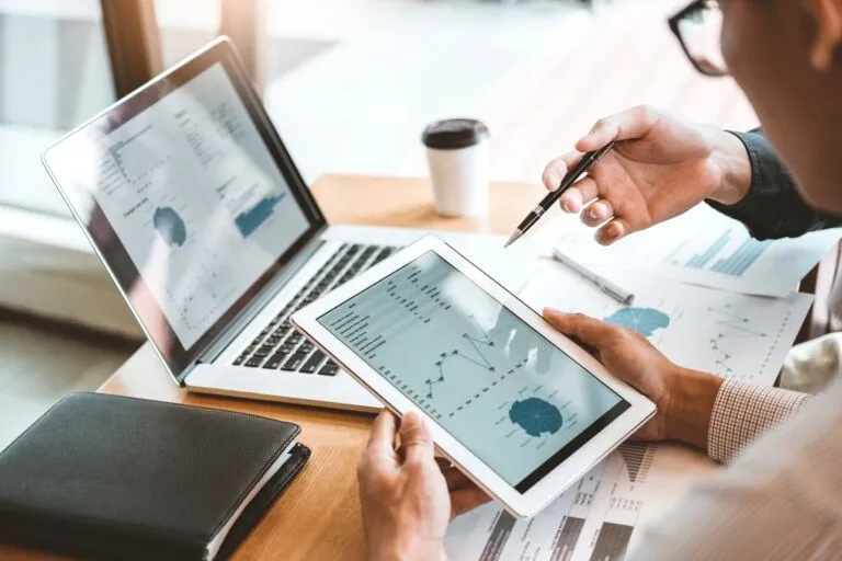 Two people conduct due diligence as they review financial charts and graphs on a tablet and laptop at a desk, surrounded by documents, a coffee cup, and a notebook.
