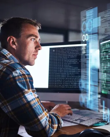 A man in a plaid shirt works at a computer with multiple screens displaying code and digital data in a dimly lit office.