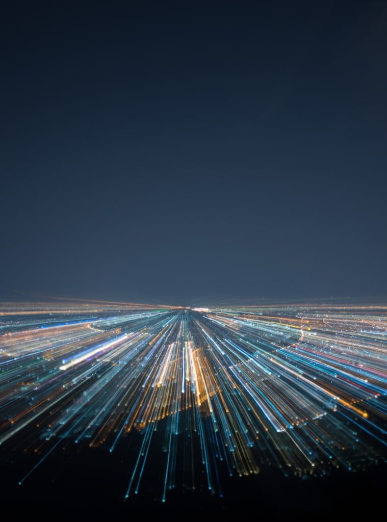 A long-exposure photo of city lights at night, creating streaks of blue, white, and yellow light that radiate outward under a dark sky.