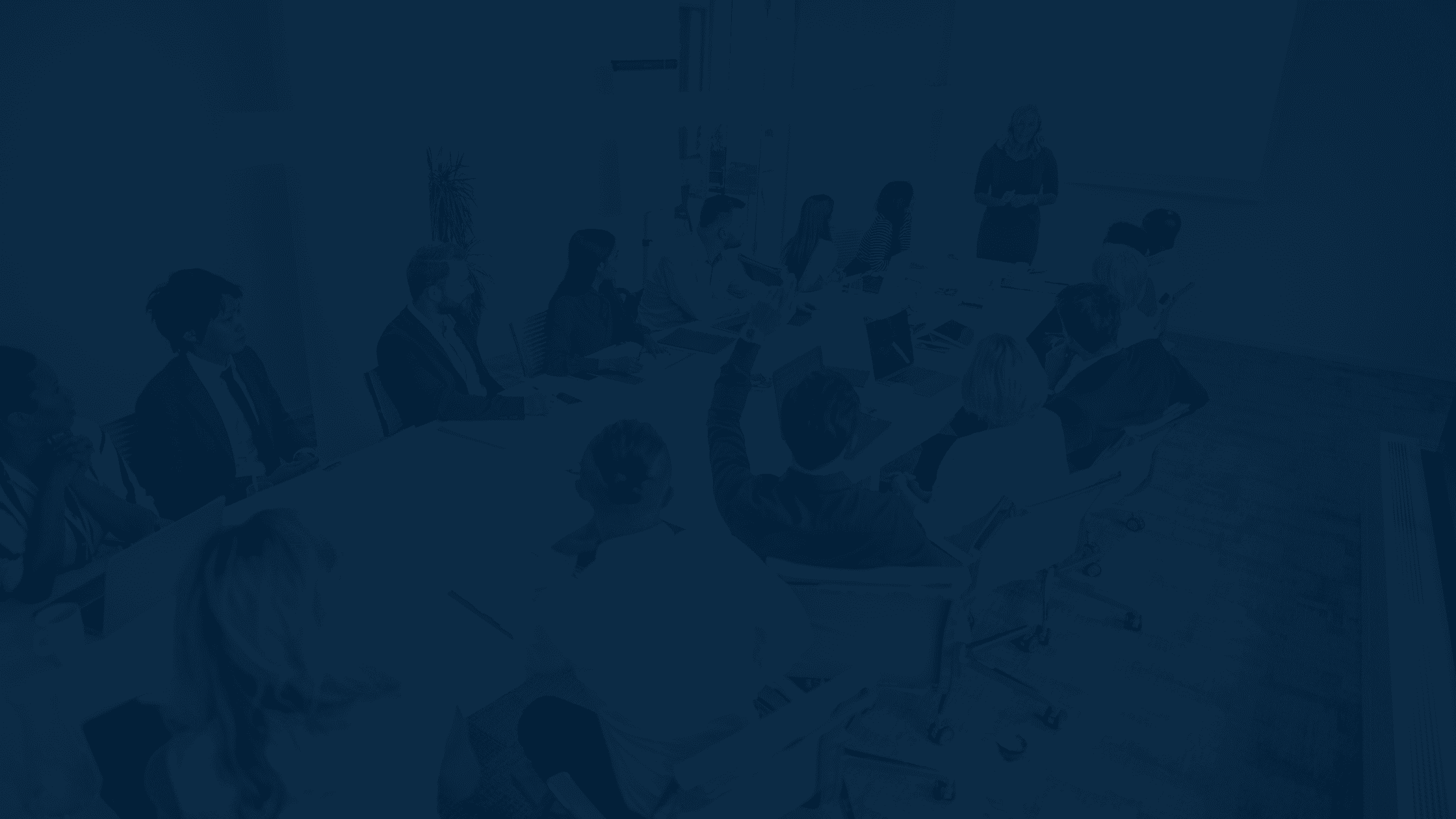 A group of people sit around a conference table while a person stands in front of a screen giving a presentation.