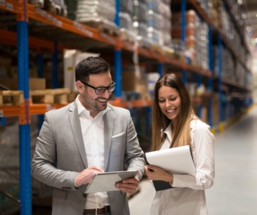 Two people stand in a warehouse aisle, smiling and looking at documents and a tablet. Shelves stocked with boxes and items are visible in the background.