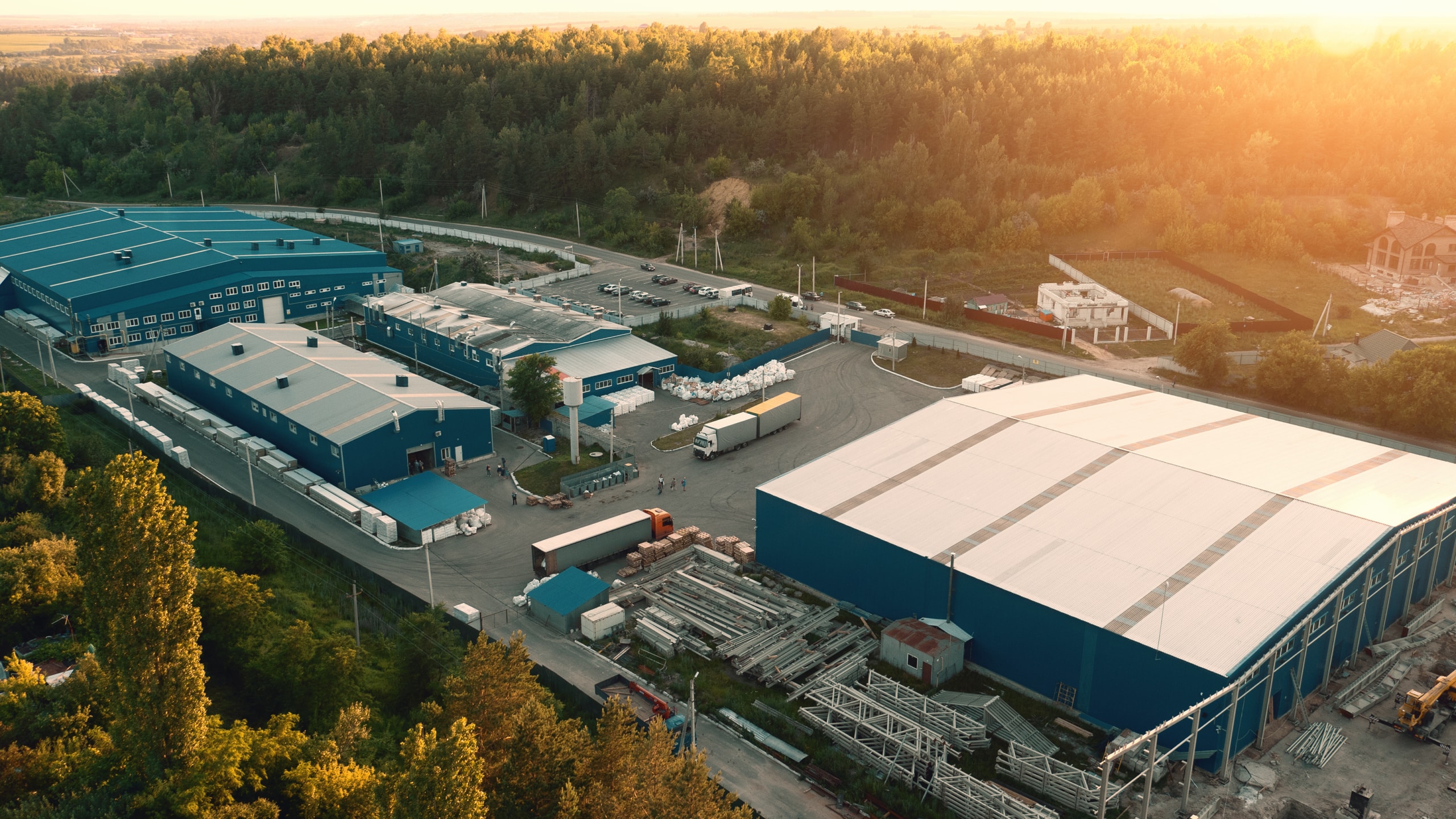 Aerial view of an industrial complex with several large blue-roofed warehouses, surrounding vehicles, and a wooded area in the background at sunset.