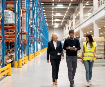 Three people walk through a warehouse; one in a suit, one with a tablet, and one in a yellow safety vest. Tall shelves with goods line the space.