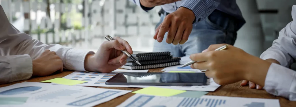 Two people hold pens and one points to a paper while discussing data on papers and tablet.