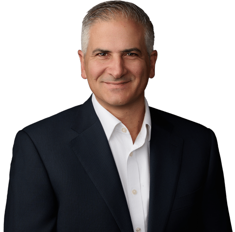 A man in a dark suit and white shirt smiles at the camera against a plain, light background.