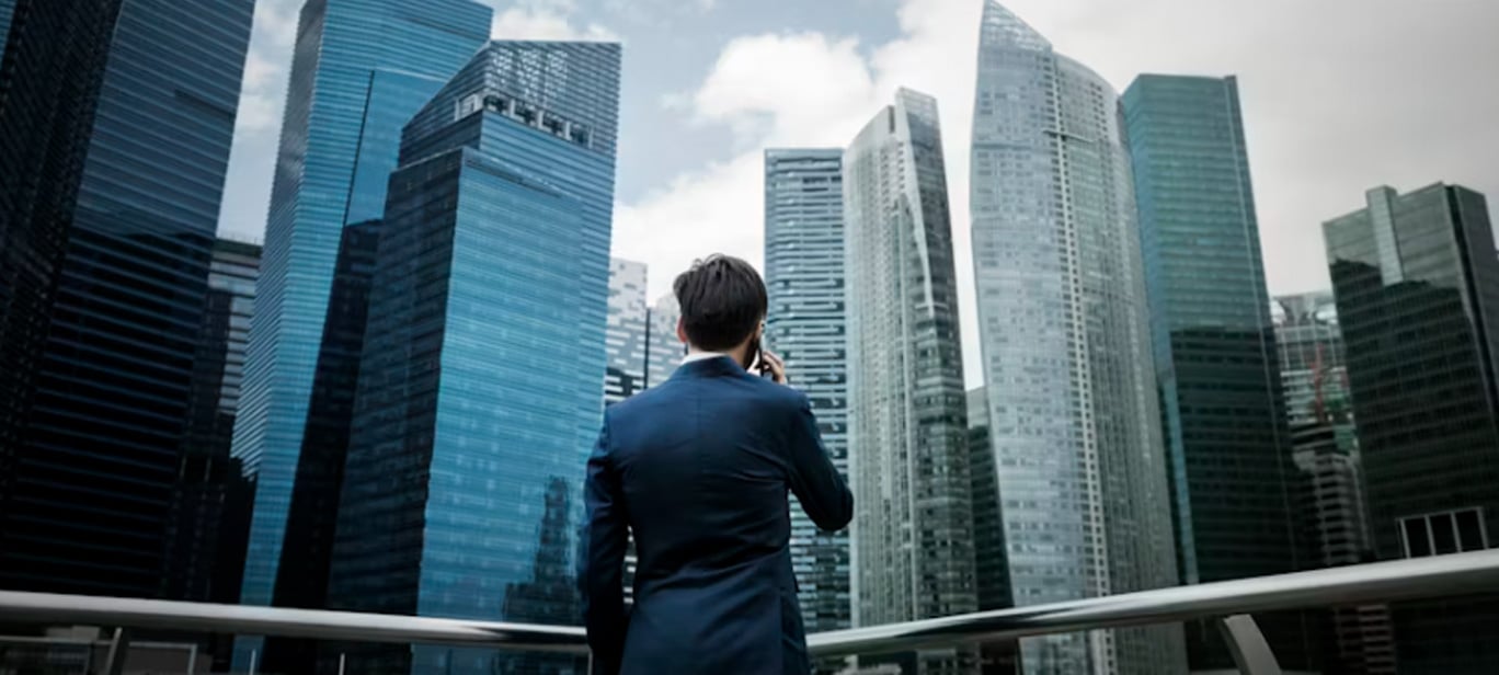 A man in a suit stands on an outdoor platform, facing and looking up at a cluster of modern glass skyscrapers in a city.
