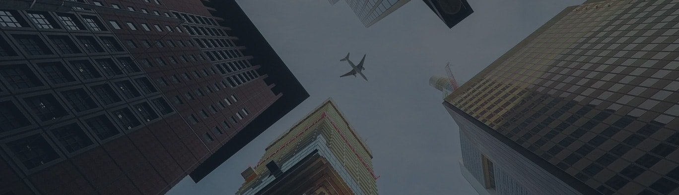 A plane flies overhead, viewed from below, surrounded by tall skyscrapers against a cloudy sky.