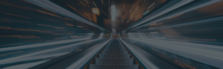 Blurred view looking up an escalator with lights and motion streaks, creating a sense of movement and speed in an indoor environment.