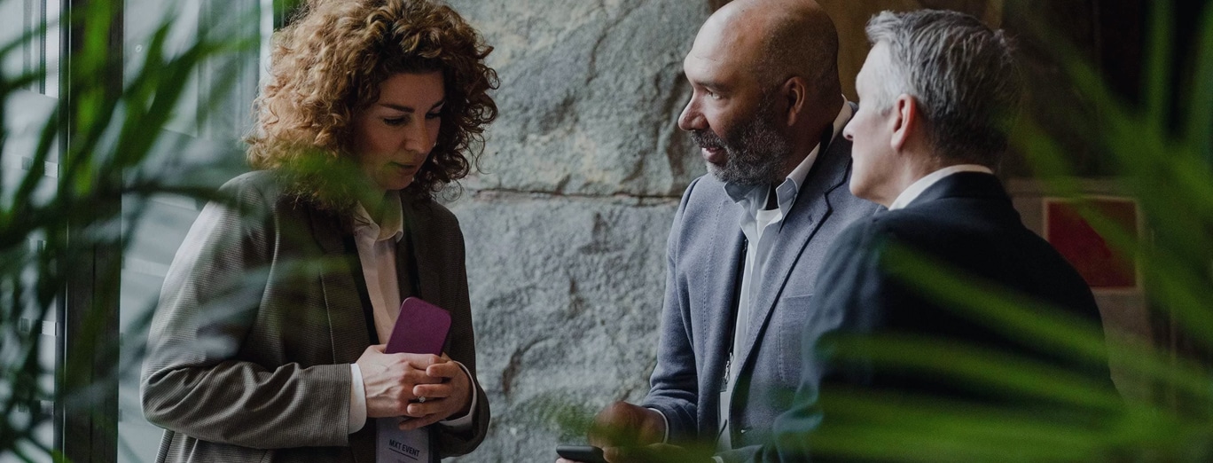 Three people in business attire stand together indoors, engaged in conversation near a window with plants visible in the foreground.
