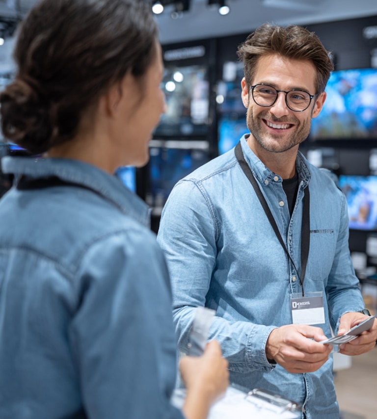 Two people wearing name tags and denim shirts converse in an electronics store, with one person smiling and holding a smartphone.